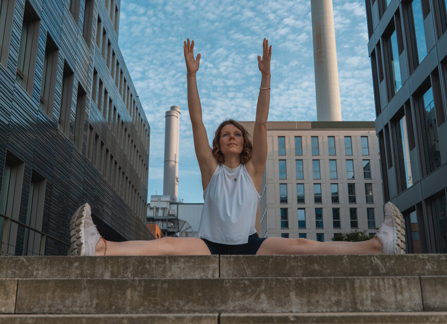 Die Yoga-Lehrerin Charlotte macht eine Yoga-Übung auf einer Treppe am Stadthafen in Münster. Im Hintergrund sind mehrere Gebäude zu sehen. | © DAV Münster